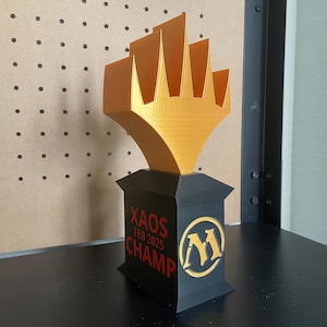 May include: A black and gold 3D printed trophy with the text "Xaos Feb 2025 Champ" and a Magic: The Gathering symbol.