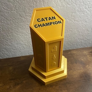 May include: A yellow trophy with the words "CATAN CHAMPION" in black lettering. The trophy has a hexagonal top and a tiered base. The sides of the trophy have embossed game icons.