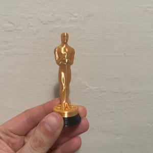 May include: A gold-coloured statuette of a man holding a sword, mounted on a black base. The figure is a replica of the iconic Oscar award, a symbol of achievement in the film industry.