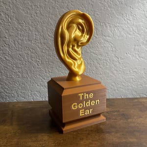 May include: A golden ear-shaped sculpture atop a brown, tiered base. The base features the text "The Golden Ear" in gold lettering. The sculpture is displayed on a wooden surface against a textured gray wall.