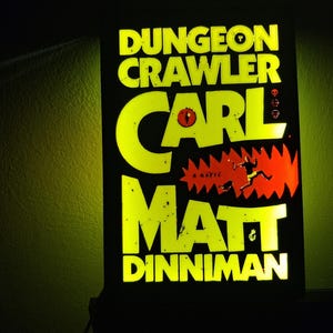 Könnte beinhalten: Ein leuchtendes Schild mit dem Text "DUNGEON CRAWLER" und dem Namen "CARL MATT DINNIMAN" in leuchtend gelben Buchstaben. Der Hintergrund ist schwarz mit einer roten und orangefarbenen Grafik eines Monsters und einer laufenden Figur.