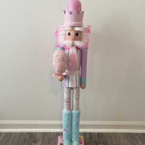Puede incluir: Figura decorativa de cascanueces en colores pastel. La figura tiene una corona rosa, una barba de piel sintética blanca y rosa, y sostiene un algodón de azúcar. Las piernas están adornadas con botas de piel sintética azul, y la base es rosa con decoraciones de caramelos.
