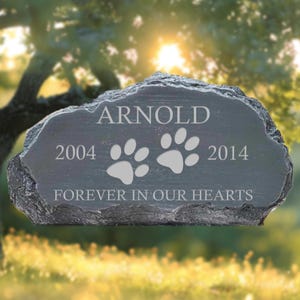 May include: A dark gray pet memorial stone with the name "ARNOLD", paw prints, and the years 2004 and 2014. The inscription "FOREVER IN OUR HEARTS" is also included. The stone has a natural, rough edge.