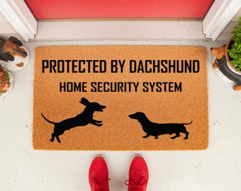 May include: A rectangular tan coir doormat featuring the black text PROTECTED BY DACHSHUND HOME SECURITY SYSTEM and two black silhouettes of dachshund dogs, one jumping and one standing, centered on the mat surface.