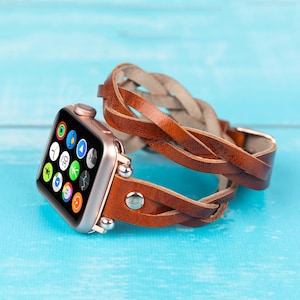 May include: A rose gold Apple Watch with a brown braided leather band. The watch face displays colorful app icons. The band wraps around the watch twice and is secured with a silver buckle and rivets. The background is a light blue surface.