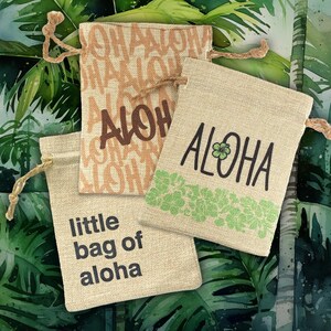 May include: Three rectangular beige drawstring pouches made of a woven fabric. One pouch features the word ALOHA in black with a green flower, another has the text little bag of aloha in black, and the third has ALOHA in brown.