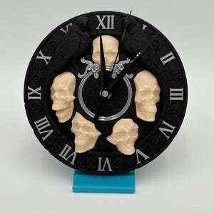 May include: A black, round clock with Roman numerals and a skull design. Two black ravens are at the top, and five skulls are placed around the clock face. The clock is on a blue stand.