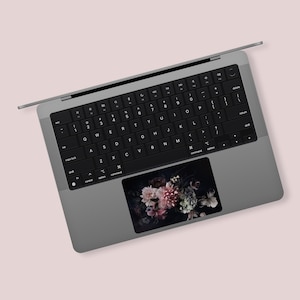 Peut inclure: Un ordinateur portable argenté avec un clavier noir et un motif floral sur le pavé tactile. Le motif floral est dans des tons de rose, blanc et vert.