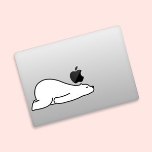 May include: A white sticker of a polar bear lying down on a silver laptop. The bear is positioned below the black Apple logo. The background is a light pink color.