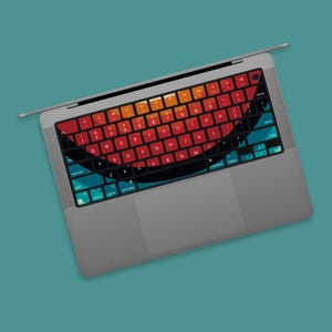 May include: A gray laptop with a colorful keyboard skin. The skin features a gradient design with red, orange, and teal hues. The keyboard keys are black with white lettering. The laptop is open on a teal background.