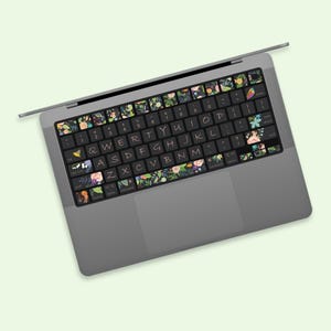 Könnte beinhalten: Ein grauer Laptop mit einem floralen Tastaturaufkleber. Der Aufkleber zeigt eine Vielzahl von Blumen, Blättern und anderen botanischen Elementen in Grün-, Rosa- und Weißtönen. Die Tasten sind schwarz mit weißer Beschriftung.