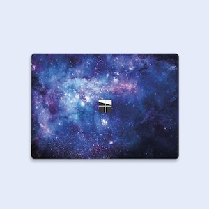 May include: A laptop skin with a galaxy design featuring a blue, purple, and black background with white stars. The skin has a cutout for the Microsoft logo.