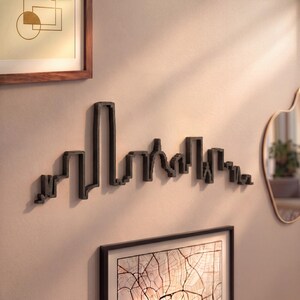 May include: A black metal wall sculpture depicting a stylized city skyline silhouette. The piece features various building heights and architectural shapes connected in a continuous line, mounted directly onto a plain, light-colored wall surface.