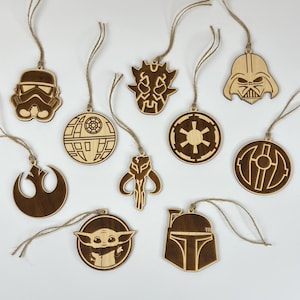 Könnte beinhalten: Eine Sammlung von Holzornamenten zum Thema Star Wars. Die Ornamente zeigen verschiedene Charaktere und Symbole, darunter Darth Vader, ein Sturmtruppler und Baby Yoda. Jedes braune Ornament hat eine Schnurschlaufe zum Aufhängen.