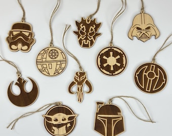 Könnte beinhalten: Eine Sammlung von Holzornamenten zum Thema Star Wars. Die Ornamente zeigen verschiedene Charaktere und Symbole, darunter Darth Vader, ein Sturmtruppler und Baby Yoda. Jedes braune Ornament hat eine Schnurschlaufe zum Aufhängen.