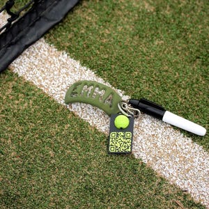 May include: An olive green curved tennis racket dampener with the name EMMA cut out, attached to a silver metal ring. A black and white marker and a black rectangular tag featuring a yellow tennis ball icon and a QR code are also attached.
