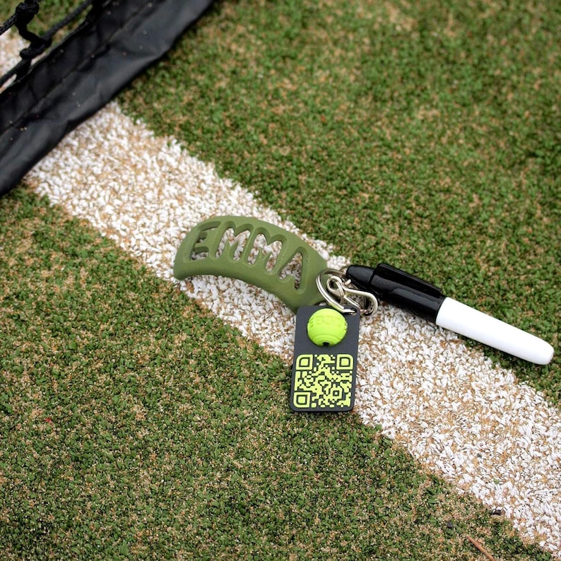May include: An olive green curved tennis racket dampener with the name EMMA cut out, attached to a silver metal ring. A black and white marker and a black rectangular tag featuring a yellow tennis ball icon and a QR code are also attached.