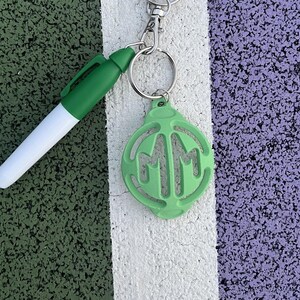 May include: A green keychain with a circle charm featuring the letters "MM" in a white outline. The keychain is attached to a silver key ring and a silver chain.