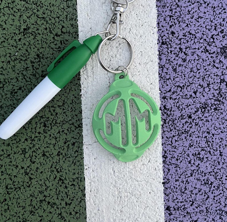 May include: A green keychain with a circle charm featuring the letters "MM" in a white outline. The keychain is attached to a silver key ring and a silver chain.