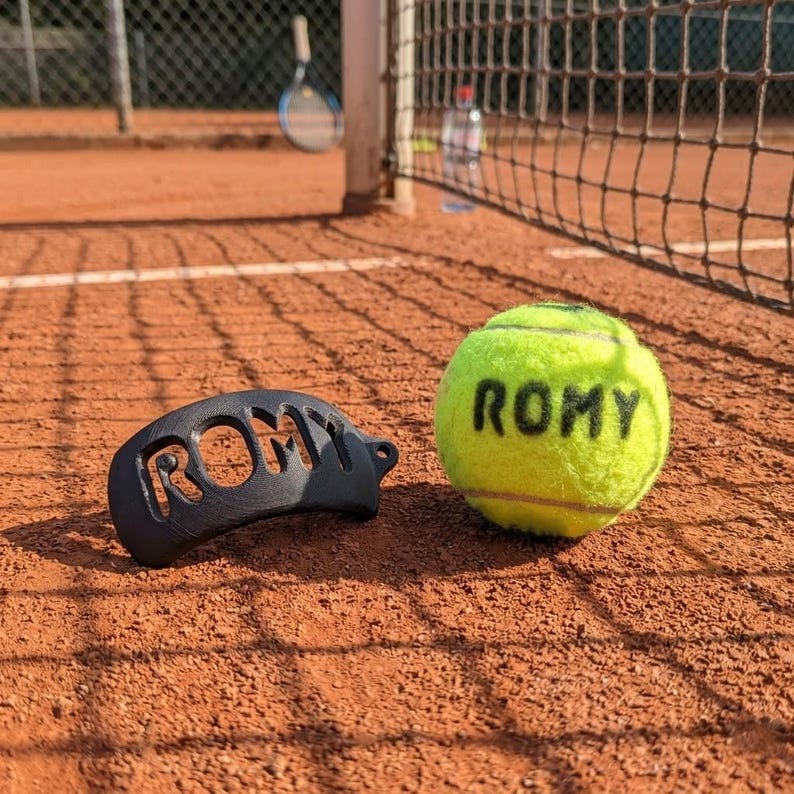 May include: A black plastic tennis ball saver with the name ROMY cut out in bold letters, positioned next to a bright yellow tennis ball with the name ROMY printed in black ink on the fuzzy surface of the ball, resting on a clay tennis court.