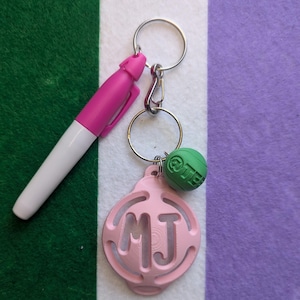 May include: A pink keychain with a silver key ring and a green charm. The keychain has a pink circle with the letters "MJ" cut out. The charm is a green sphere with the word "LIFE" printed on it.