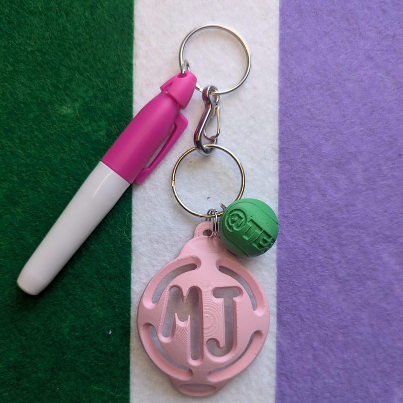 May include: A pink keychain with a silver key ring and a green charm. The keychain has a pink circle with the letters "MJ" cut out. The charm is a green sphere with the word "LIFE" printed on it.