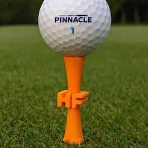 Può includere: Una pallina da golf bianca Pinnacle con il numero 1 è posizionata su un tee da golf arancione. Il tee presenta le lettere "HF". Lo sfondo è erba verde, che suggerisce un campo da golf.