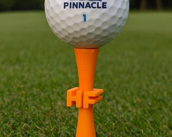 Può includere: Una pallina da golf bianca Pinnacle con il numero 1 è posizionata su un tee da golf arancione. Il tee presenta le lettere "HF". Lo sfondo è erba verde, che suggerisce un campo da golf.