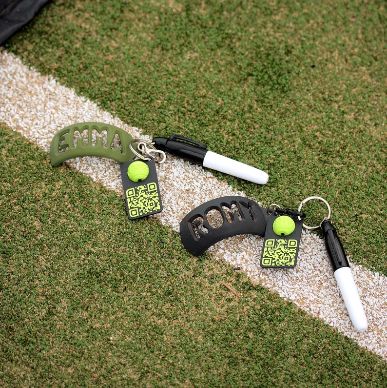 May include: Two keychains featuring curved olive green and black silicone pieces with the names EMMA and ROMY cut out. Each includes a small black marker and a rectangular black tag with a bright green tennis ball icon and a QR code.