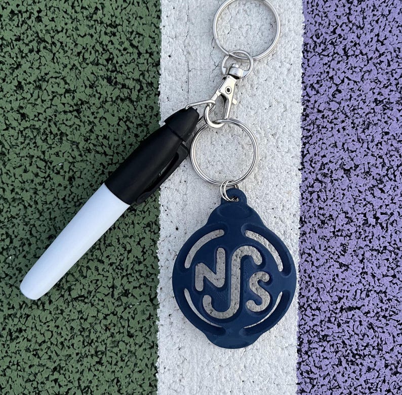 May include: A blue keychain with the letters "NS" in a circle design. The keychain is attached to a silver key ring and a white dry-erase marker.