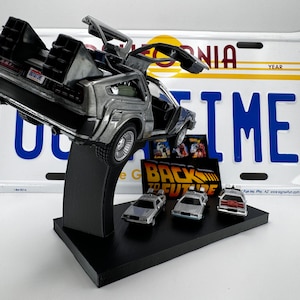 May include: A silver DeLorean time machine from the Back to the Future movie franchise, with the doors open and the flux capacitor visible. The car is on a black stand with a 'Back to the Future' logo and four smaller DeLorean models.