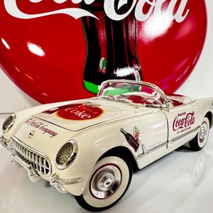 May include: White 1957 Chevrolet Corvette convertible model car with Coca-Cola branding. The car has a red and white Coca-Cola logo on the hood and the words "Drink Coca-Cola" on the side. The car is sitting on a white surface.
