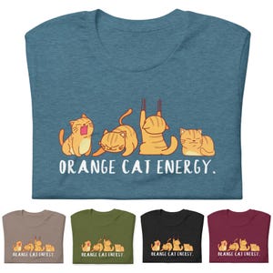 Puede incluir: Una camiseta azul con un gráfico de cinco gatos naranjas en diferentes poses. El texto "ORANGE CAT ENERGY." está impreso debajo de los gatos.