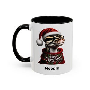 May include: A white ceramic mug with a black handle and interior rim. The mug features a graphic of a snake wearing a red Santa hat and a red sweater with the text Merry Christmas. The word Noodle is printed in black below the graphic.