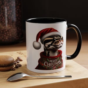 May include: A white ceramic mug with a black interior and handle, featuring a graphic of a snake wearing a red Santa hat and a red sweater with the text Merry Christmas printed in white, surrounded by a decorative wreath of holly and berries.