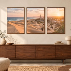 May include: A triptych of framed wall art prints featuring a coastal landscape with sand dunes, a lighthouse, and a sunset over the ocean. The three vertical rectangular frames are hung side-by-side above a long, dark wood media console cabinet.