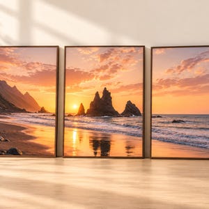 May include: A triptych wall art set featuring a coastal landscape at sunset. The three framed prints display jagged rock formations rising from the ocean, a sandy beach, and a vibrant orange sky with clouds, all enclosed in thin dark brown frames.