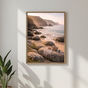 Può includere: Stampa incorniciata di una scena di spiaggia con sabbia, rocce e un mare calmo. Un cappello di paglia, una coperta e un cestino sono appoggiati su una roccia. Il cielo è rosa e le montagne sullo sfondo.