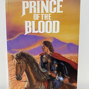 May include: A book cover with a fantasy illustration of a man on horseback riding through a desert landscape. The title of the book is "Prince of the Blood" by Raymond E. Feist. The book is part of the Riftwar Saga.