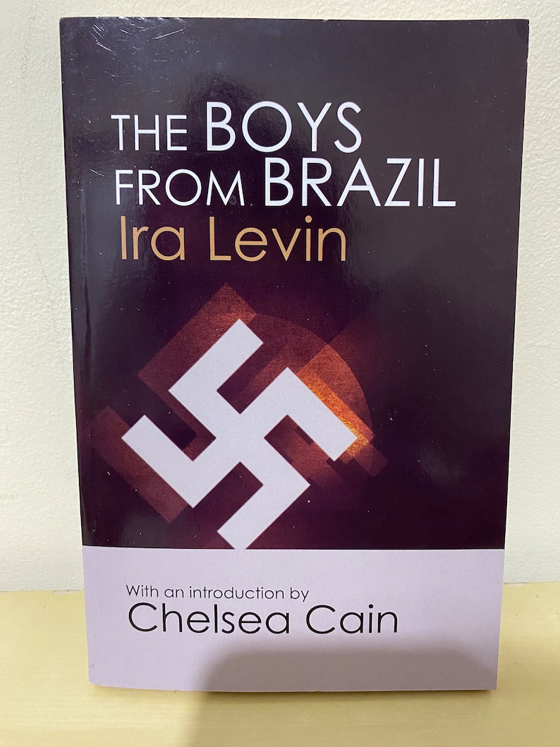 May include: A book cover with a black background and white text that reads "The Boys From Brazil" by Ira Levin. The cover also features a white swastika symbol and the text "With an introduction by Chelsea Cain".