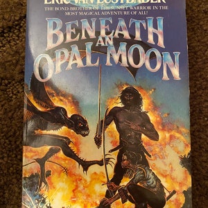 Puede incluir: Una portada de novela fantástica de bolsillo con fondo azul y el título "Beneath an Opal Moon" en letras blancas. La portada presenta a un hombre de piel oscura con una espada, una criatura con alas y una mujer con una camisa azul y pantalones negros.