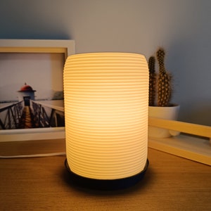 May include: A white ceramic table lamp with a ribbed design and a black base. The lamp is switched on and emitting a warm, yellow light.