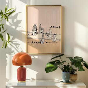 May include: A framed print with a peach background featuring a line of black birds and several white hands in various hand gestures. The print is hanging on a white wall with a brown lamp and a green plant in a blue pot on a white table.