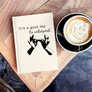 Puede incluir: Un cuaderno color crema con el texto "It's a good day to interpret" y una ilustración negra de manos que firman. Una taza de café con arte latte está sobre un platillo negro junto al cuaderno.