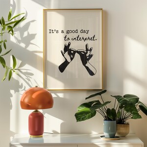 May include: A framed art print with the text "It's a good day to interpret" and a black and white hand gesture illustration. A red and gold table lamp and potted plants are on a white surface.