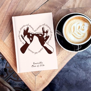 Puede incluir: Un cuaderno crema con una ilustración marrón de manos formando un corazón, rodeado por el texto "interpreter asl". El cuaderno también tiene el nombre "Esmeralda" y "Class of 2026" impresos. Una taza de café está al lado.