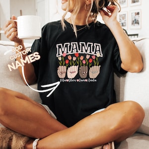 May include: Black t-shirt with the word "MAMA" in pink and white letters.  The shirt features a design of hands in different skin tones holding pink and white flowers.  The text "Mia Oliver Liam Amelia" is printed below the hands.