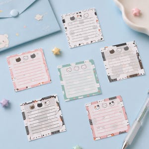 Könnte beinhalten: Eine Sammlung von Haftnotizen mit Katzenmotiven in verschiedenen Farben, darunter Rosa, Grün und Weiß, mit linierten Bereichen zum Schreiben. Jede Notiz hat einen Rand mit Katzenillustrationen und eine Kopfzeile mit drei Katzenköpfen. Ein hellblauer Umschlag und sternförmige Dekorationen sind ebenfalls zu sehen.