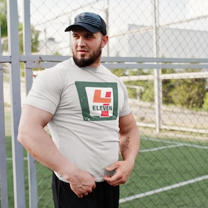 Op de afbeelding: Een wit T-shirt met een groen, rood en oranje logo met de tekst "7-Eleven". De persoon die het shirt draagt staat voor een hek van gaas.