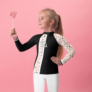 Könnte beinhalten: Ein langärmliges Shirt in Schwarz und Weiß mit einem rosa und weißen Sushi-Muster auf dem rechten Ärmel. Das Shirt hat den Text "Let's Roll" auf der Vorderseite aufgedruckt. Ein junges Mädchen trägt das Shirt und hält einen roten und weißen Lolli.